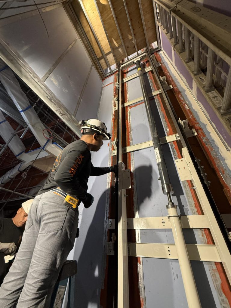 Worker in a hard hat installs elevator guide rails up a multi-story shaft.